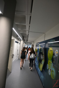 Schüler:innen laufen durch den Gang eines Universitätsgebäudes. Rechts ist eine Pinnwand zu Topologischen Isolationen zu sehen. Links sind Säulen und offene bzw. geschlossene weiße Türen.